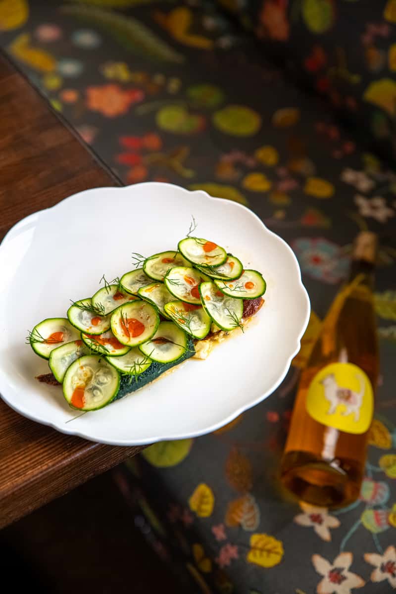 Toasted tartine topped with thin cucumber slices, dill, and hot sauce, paired with a bottle of chilled natural wine.