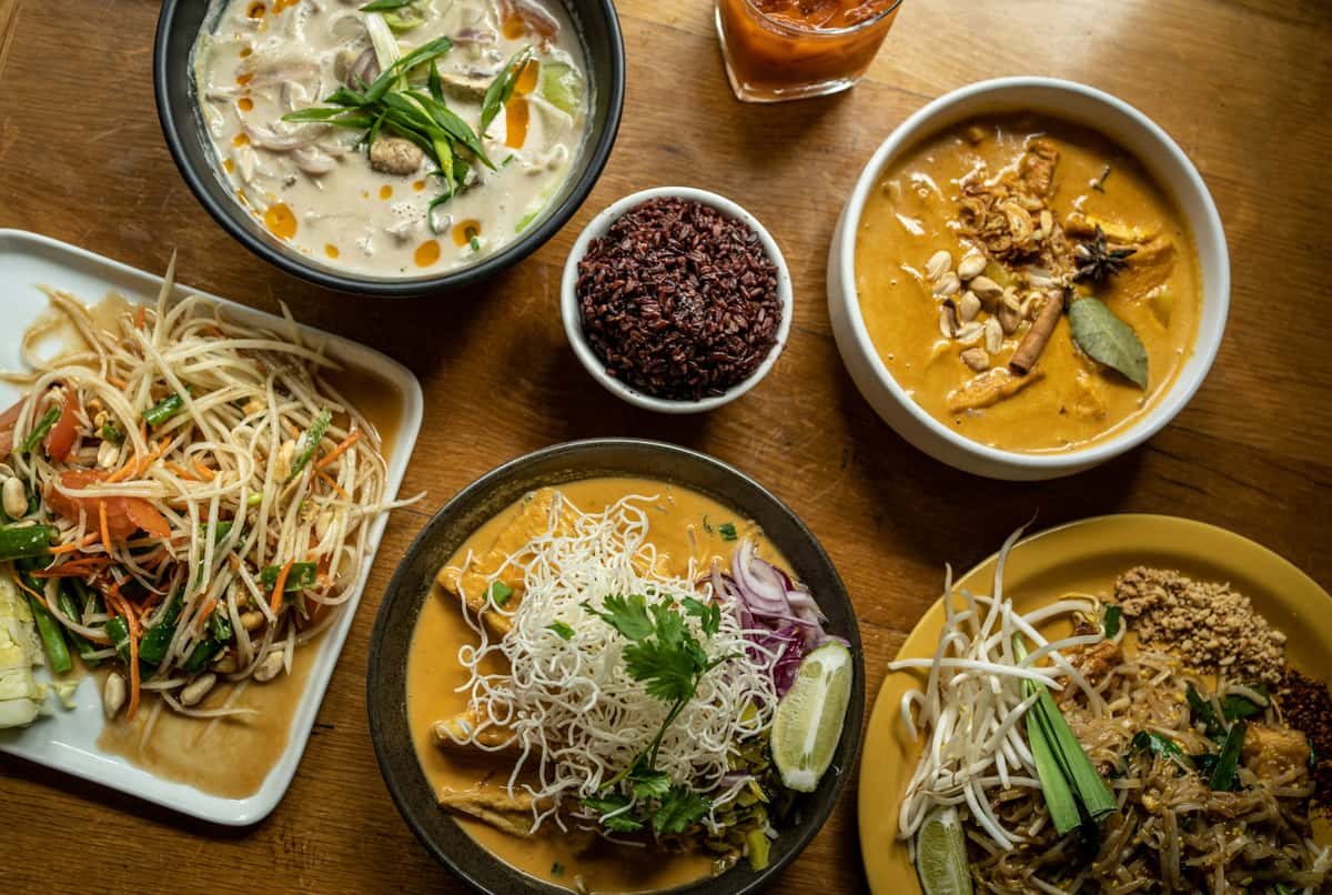 Assorted Thai dishes on a wooden table including papaya salad, curries, noodles, black rice, and pad thai with peanuts and lime.