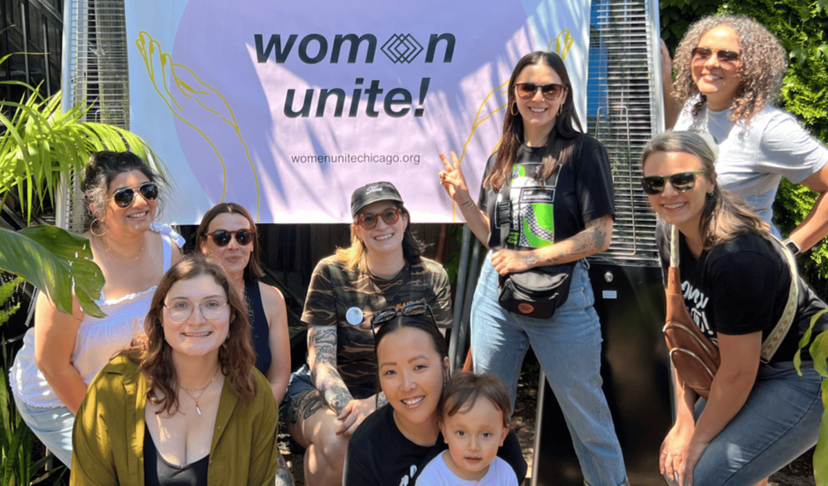 a group of women standing in front of a banner with a pink circle with the the Women Unite! logo