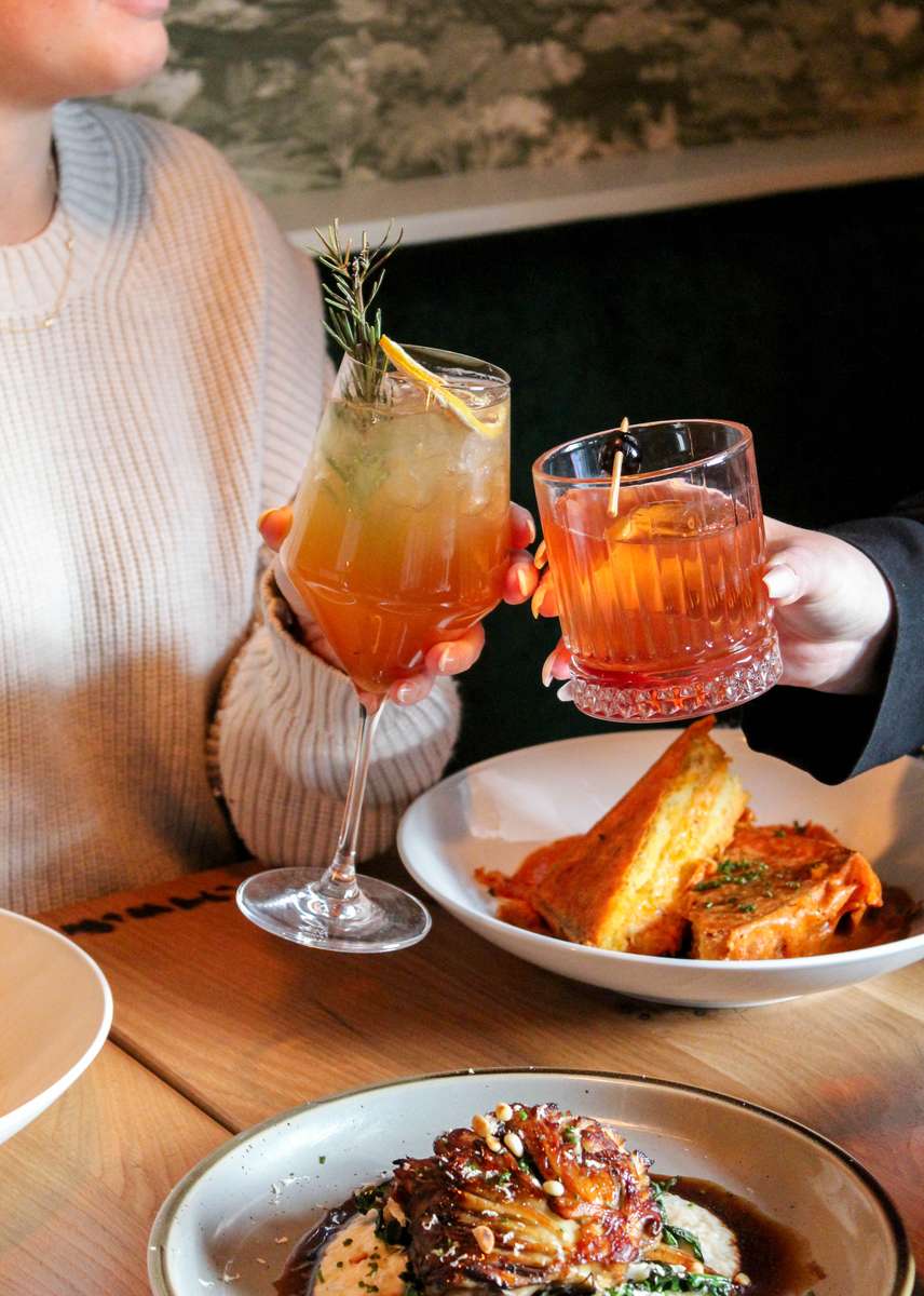 Two glasses with orange liquid cheers over a plate of grilled cheese