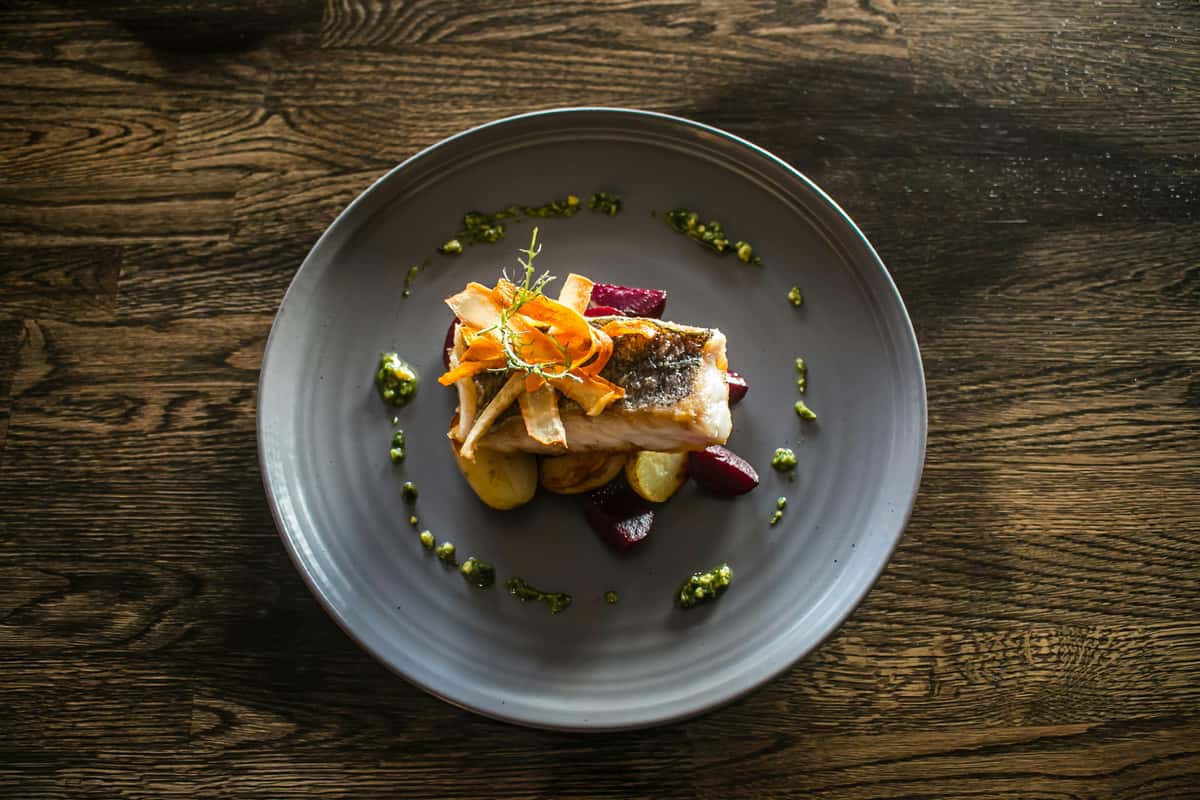 Pan-seared fish fillet topped with crispy vegetable strips, served over roasted beets and potatoes with herb sauce garnish.