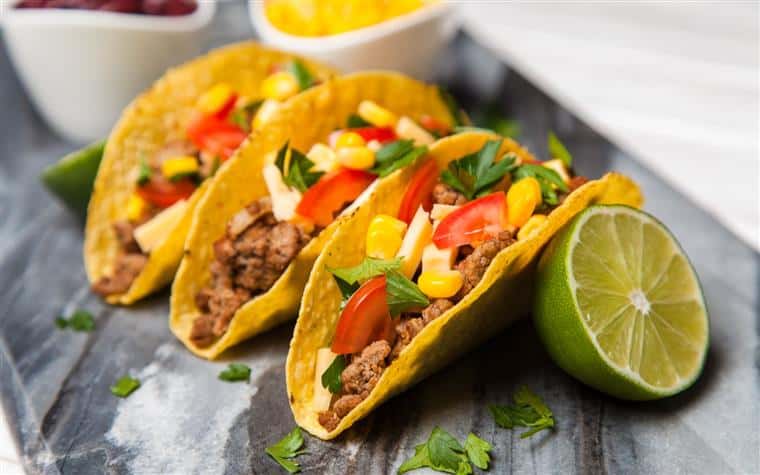 Beef tacos in a hard shell with tomatoes and corn on a slate top with lime