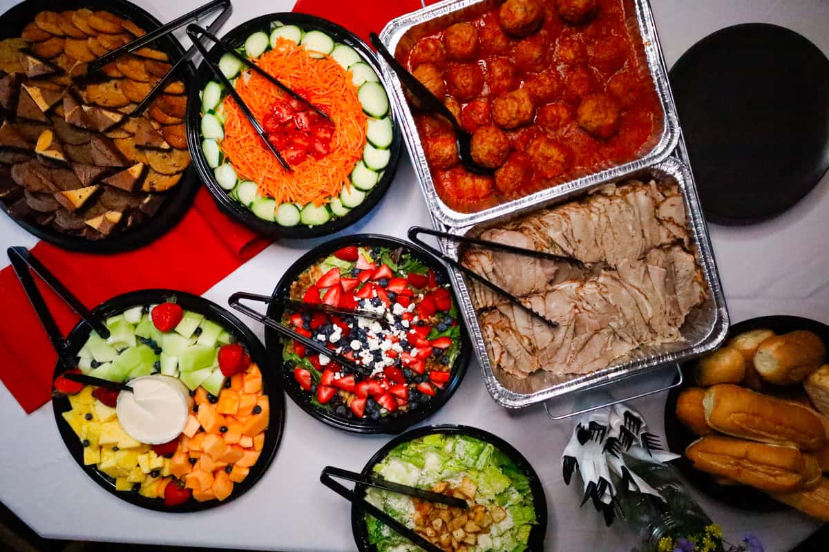 A vibrant catering spread featuring assorted meats, fresh salads, and colorful fruit platters, perfect for events. Includes meatballs, sliced turkey, and various sides.