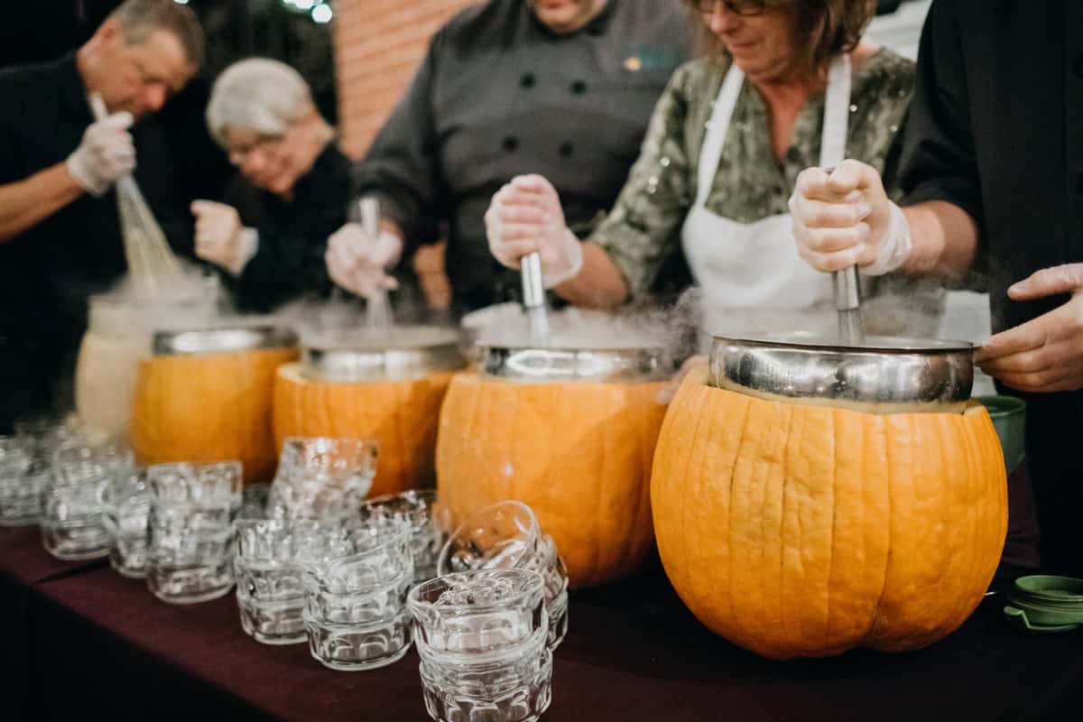 Caterers prepare a unique pumpkin soup in hollowed-out pumpkins, with steam rising, while guests enjoy the festive atmosphere, showcasing creativity in event catering.