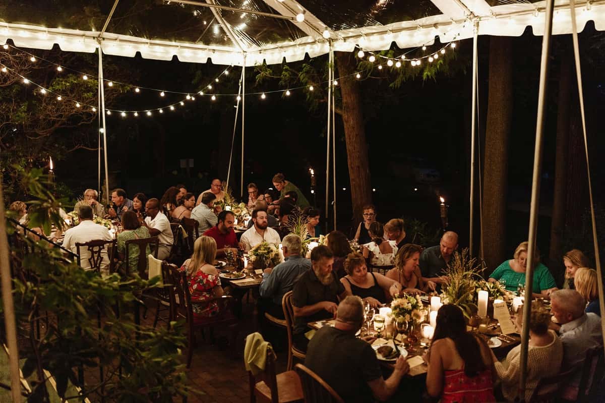 A beautifully decorated outdoor dining setup under a tent, featuring guests enjoying a meal with soft lighting, floral centerpieces, and a warm, inviting atmosphere for a special event.