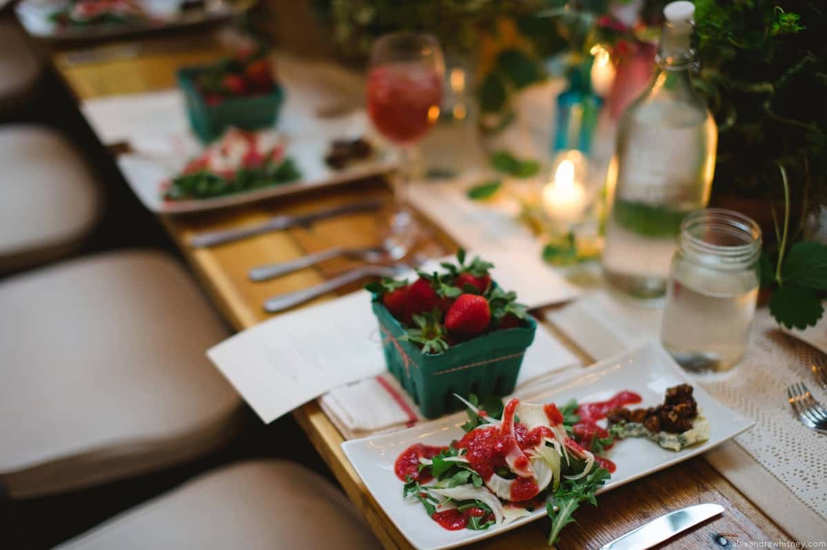 A beautifully set wedding table featuring a fresh salad with strawberries and a side of greens, accompanied by a basket of strawberries, elegant glassware, and soft candlelight.