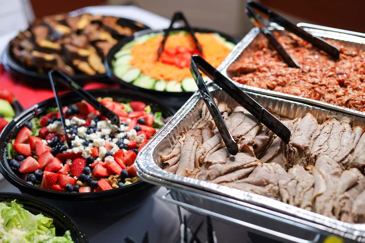 A vibrant catering spread features sliced roast beef, a fresh berry salad, and colorful vegetable platters, at a festive event.