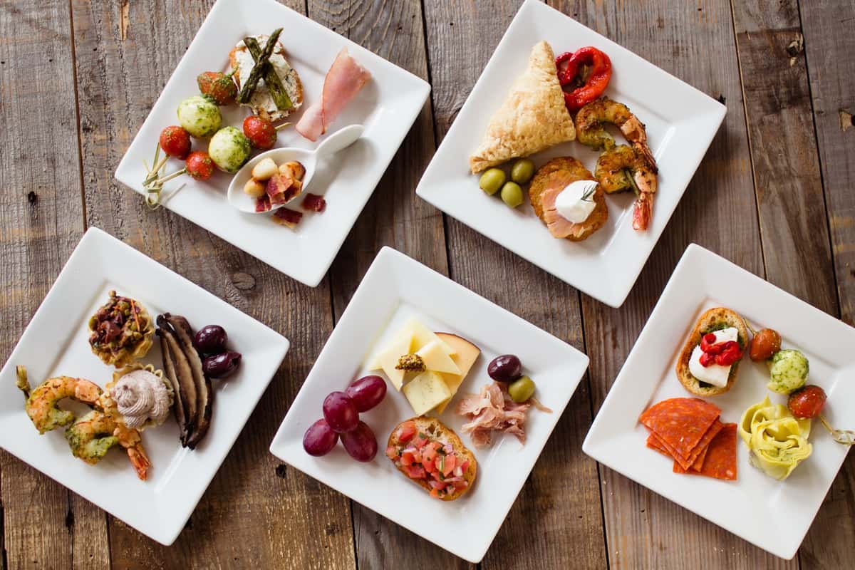 An elegant wedding catering display featuring five square plates with gourmet appetizers, including shrimp, olives, and pastries, arranged on a rustic wooden table.
