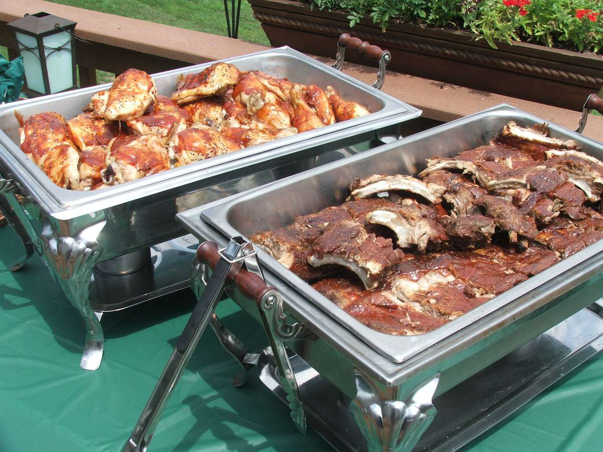 Two chafing dishes filled with delicious catering options: one with glazed chicken pieces and the other with tender ribs, set on a green tablecloth, perfect for outdoor events.