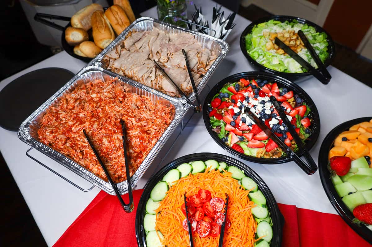 A vibrant catering spread featuring pulled chicken, sliced turkey, fresh salads with strawberries and blueberries, and assorted bread rolls, perfect for any event.