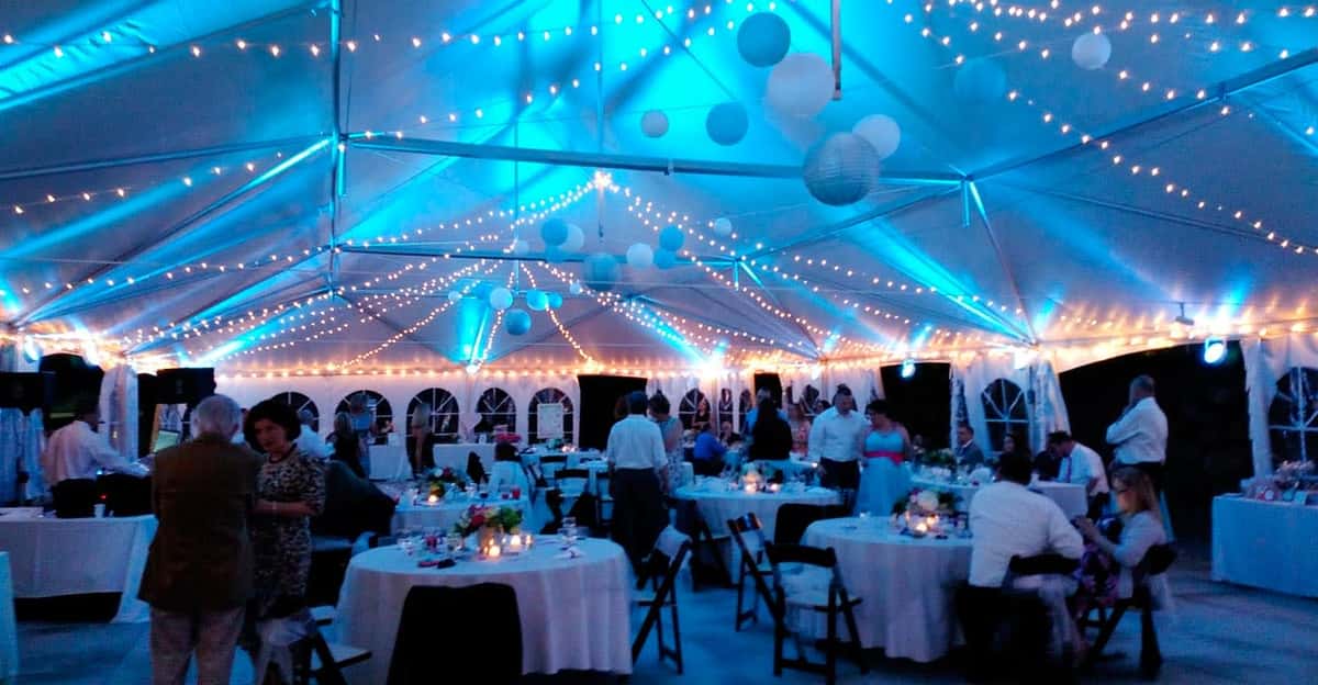A beautifully decorated event space featuring round tables set with white linens and elegant centerpieces. The ceiling is adorned with glowing string lights and soft blue uplighting, creating a warm and inviting ambiance—perfect for weddings, banquets, or special celebrations.