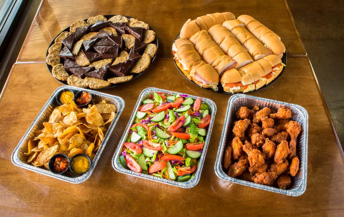 catering spread of subs, salads, wings, chips, and dessert