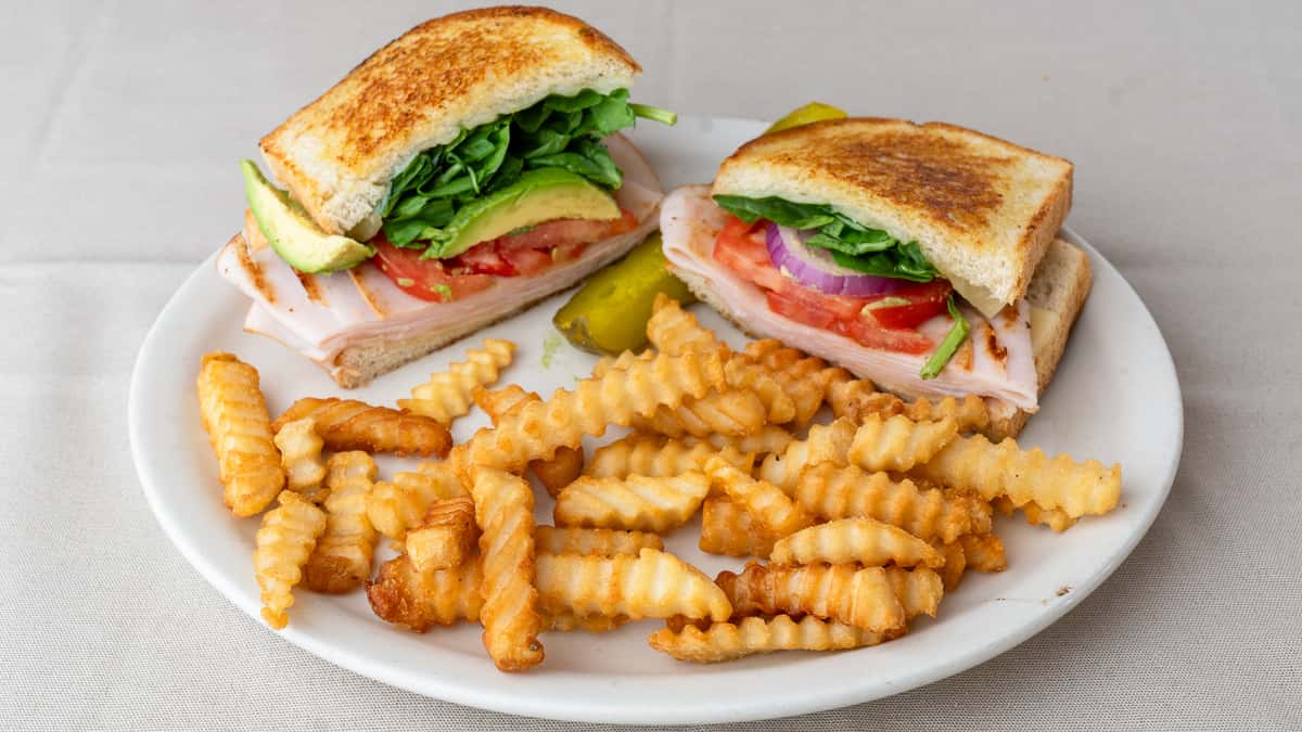 Grilled turkey sandwich with avocado, spinach, tomato, and red onion, served with crinkle-cut fries and pickle slices.