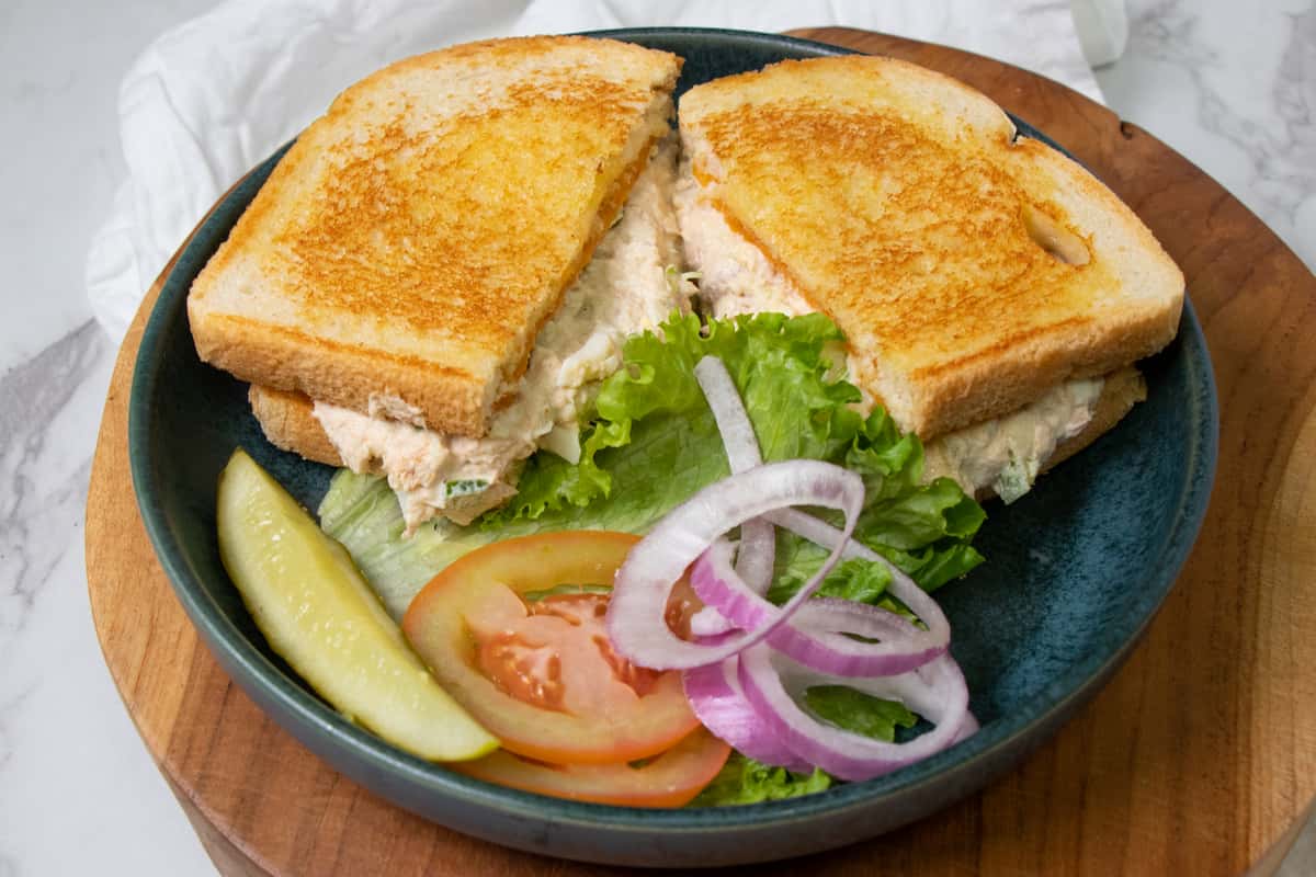 Grilled tuna salad sandwich on toasted bread, served with lettuce, tomato, red onion, and a dill pickle spear on a blue plate.