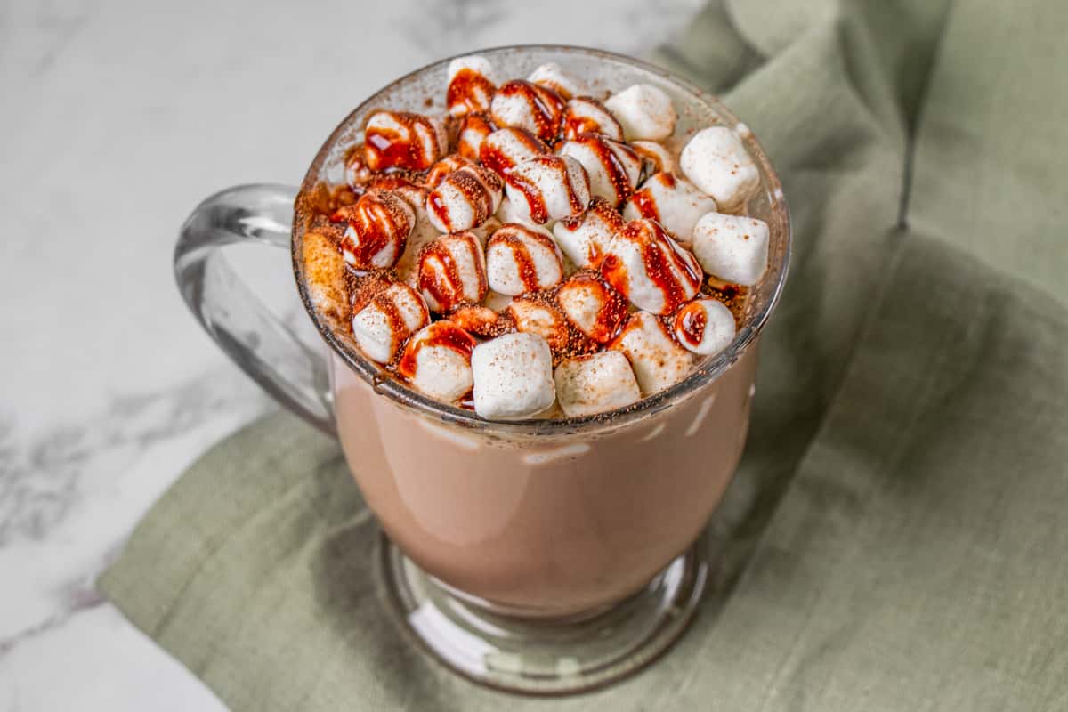 Glass mug of hot chocolate topped with mini marshmallows, cocoa powder, and chocolate drizzle on a green cloth.