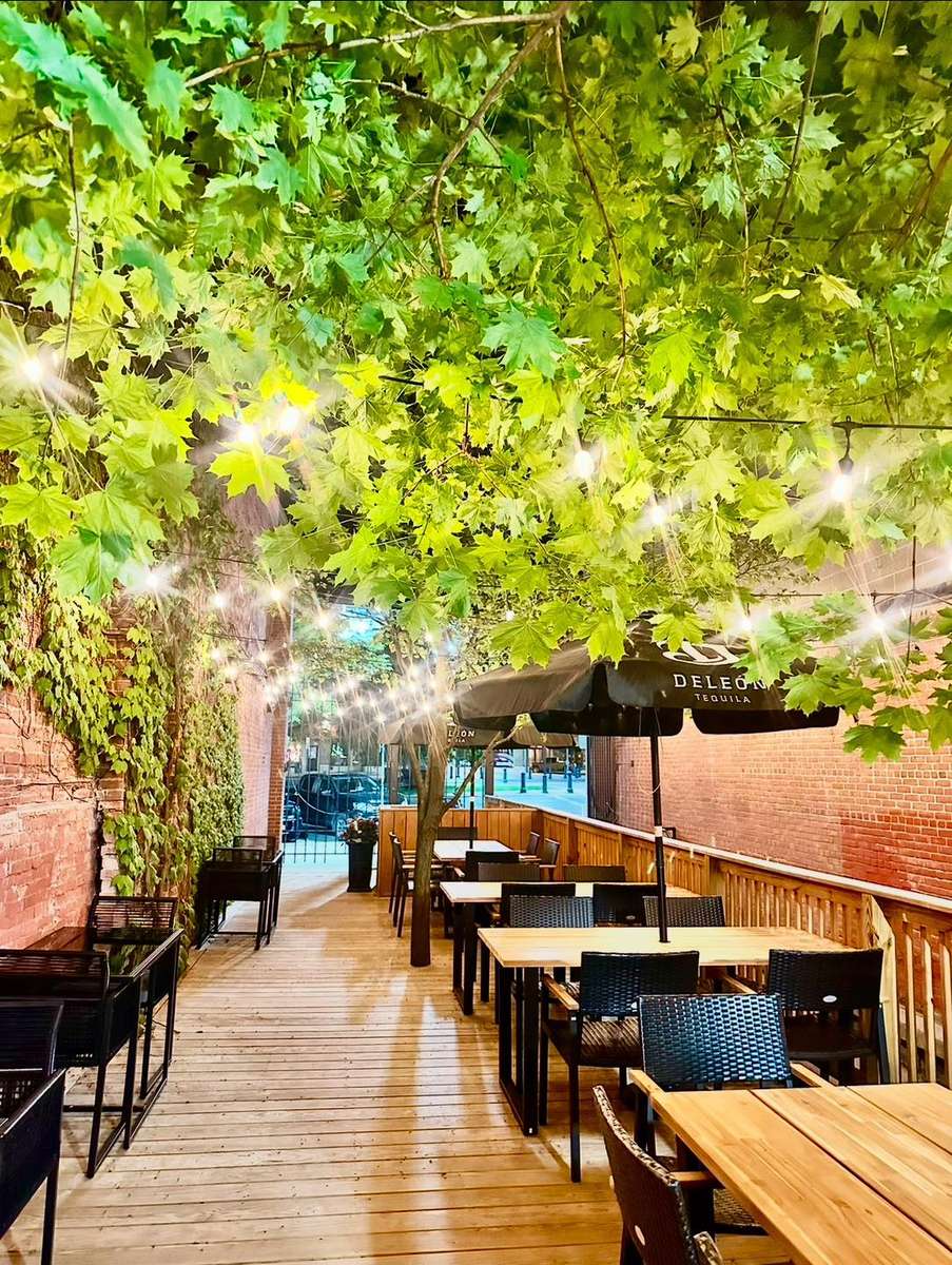 Charming outdoor patio with wooden decking, black tables, leafy overhead canopy, string lights, and a DeLeón Tequila umbrella.