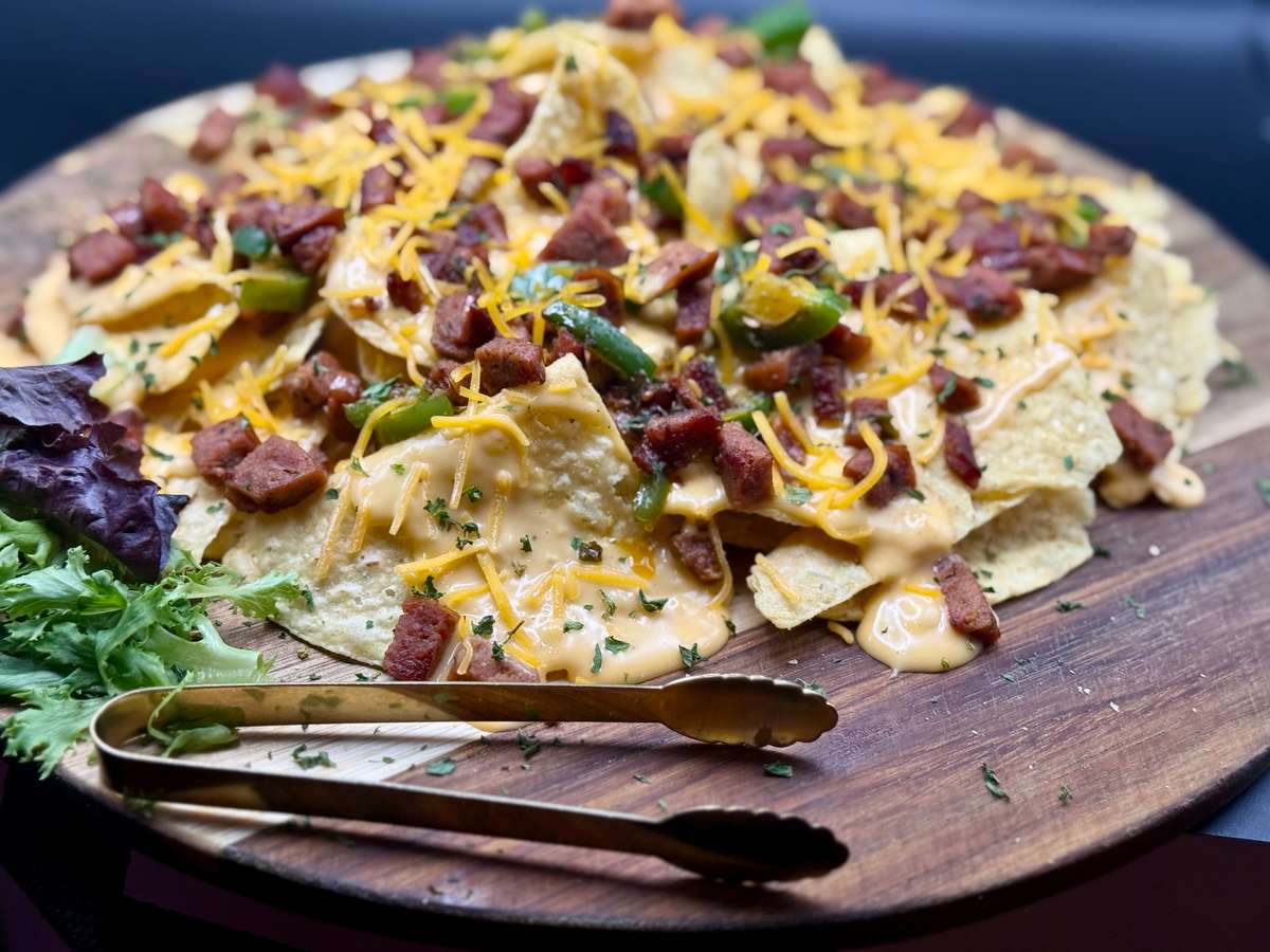 Loaded nachos topped with melted cheese, diced sausage, jalapeños, and herbs on a wooden serving board with salad greens.