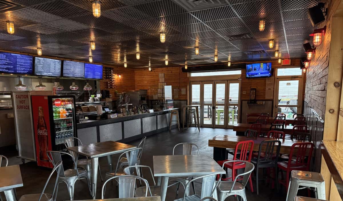 The interior of Main Street Eats shows empty tables and chairs awaiting customers beneath industrial lighting and a Coca-Cola refrigerator.