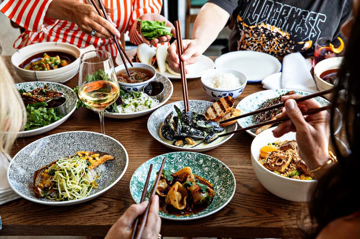 Several pair of hands using chopsticks to pickup food from family style dishes