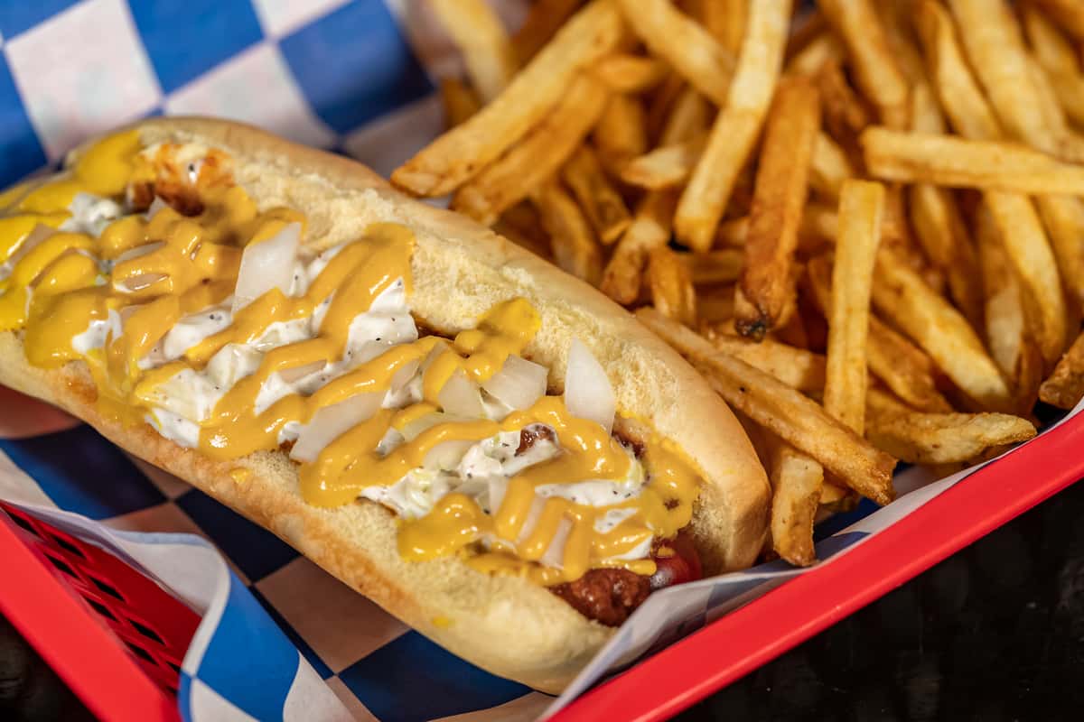 Loaded hot dog topped with chopped onions, creamy relish, and a drizzle of yellow mustard, served with a generous portion of crispy golden French fries in a red diner-style basket with checkered paper — classic American fast food presentation.