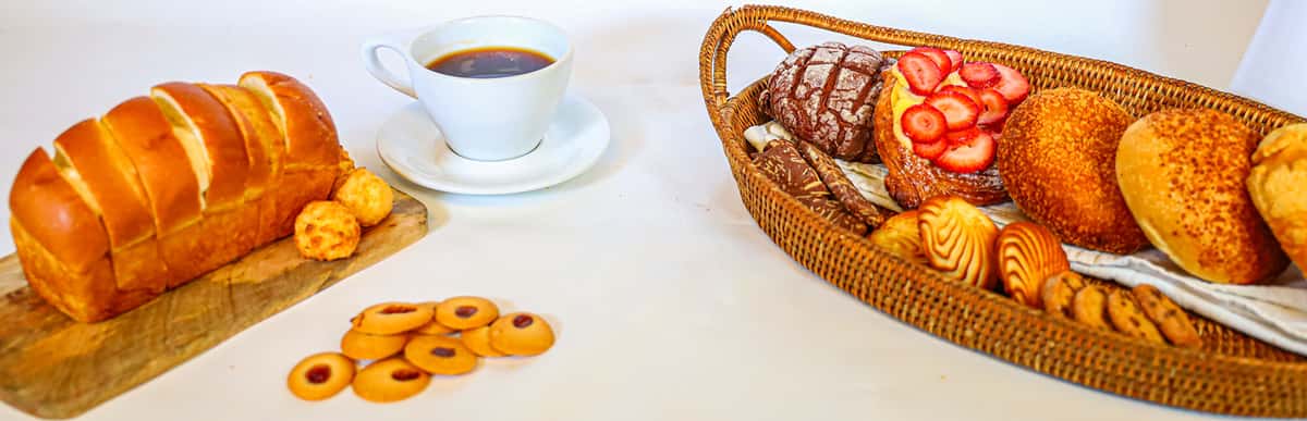 Assorted pastries and bread in basket with coffee cup, including strawberry pastry, sweet buns, and sliced loaf