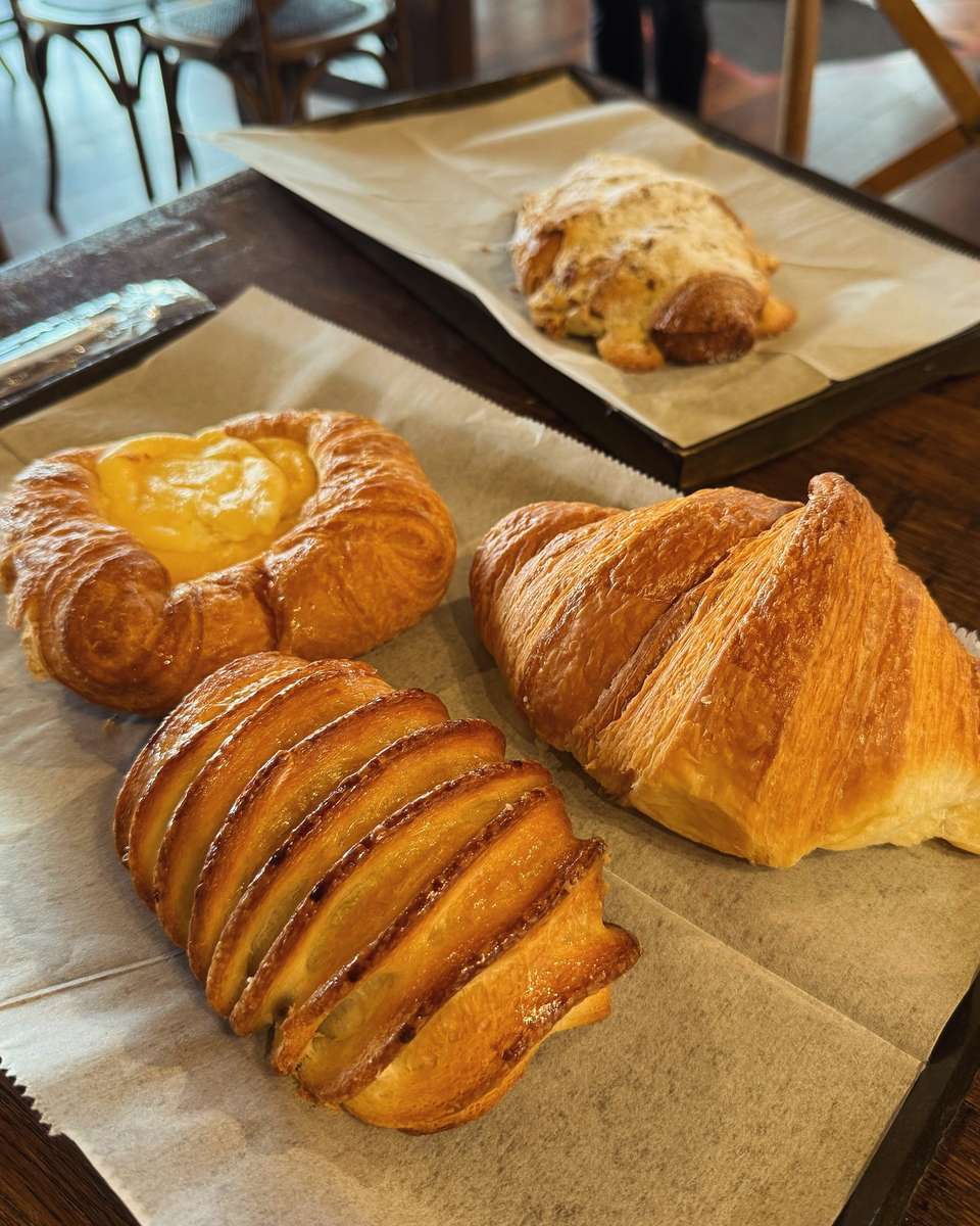 Assorted pastries on parchment-lined trays, including croissants, custard Danish, and glazed sweet bread in café setting