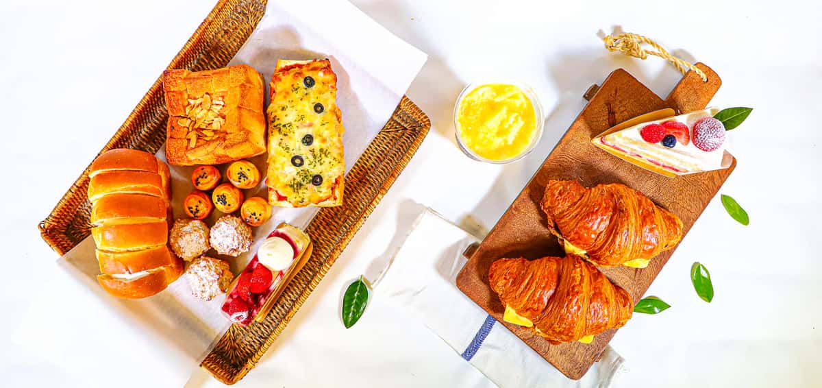 Overhead spread of assorted breads, croissants, cake slice, and pastries on wicker and wooden trays with juice