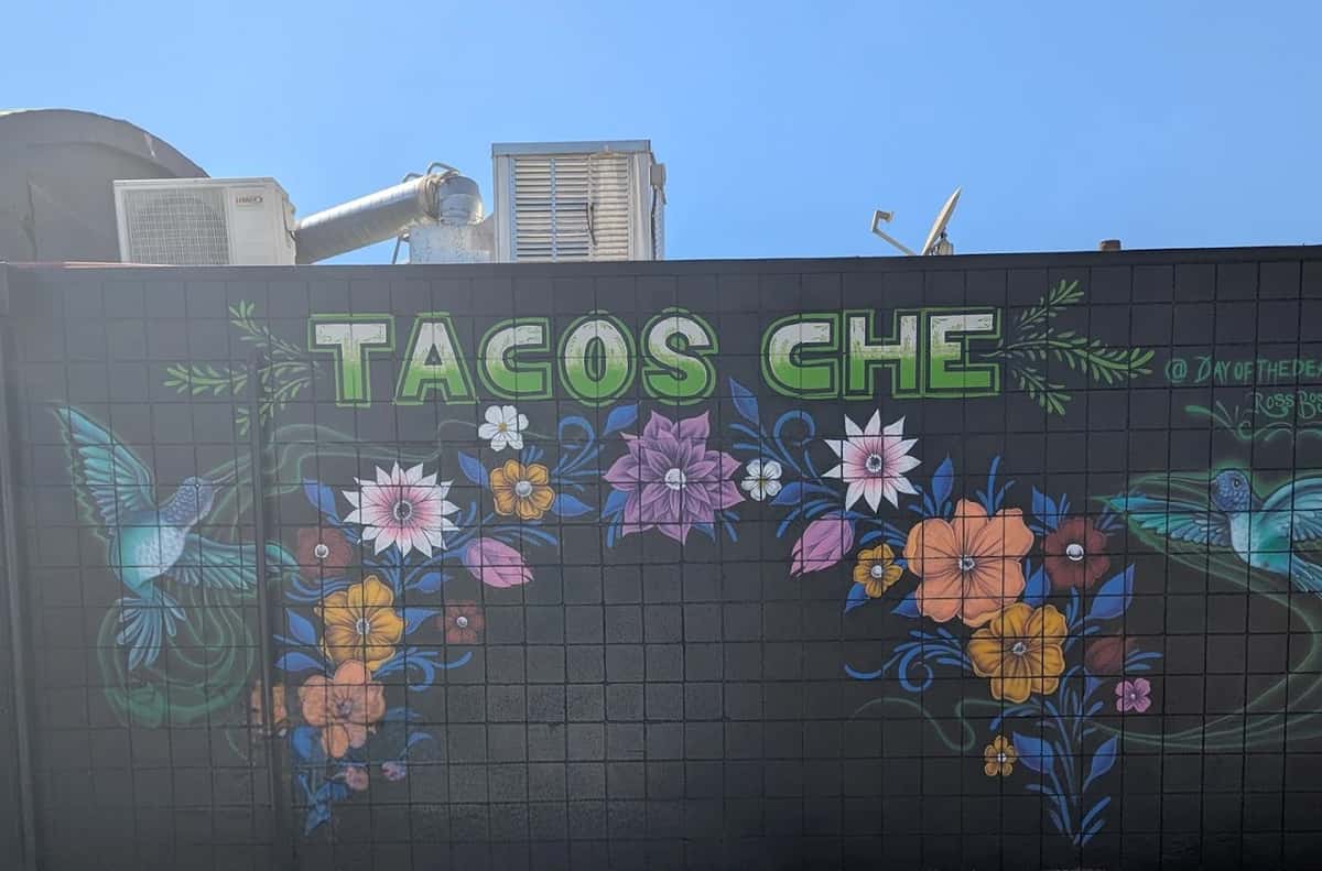 Colorful Tacos Che mural featuring vibrant floral artwork and hummingbirds on a black exterior wall under a clear blue sky.