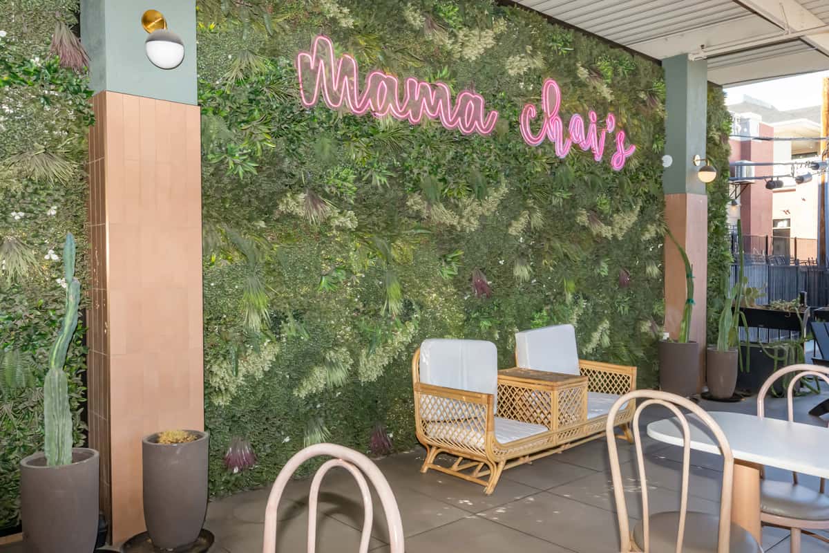 Trendy outdoor seating area at Mama Chai's, featuring modern wicker lounge chairs with white cushions, a wooden table, and a lush green plant wall with a neon pink sign. The chic café patio is accented with potted cacti and natural light, creating a vibrant, Instagram-worthy atmosphere ideal for casual dining or sipping bubble tea.