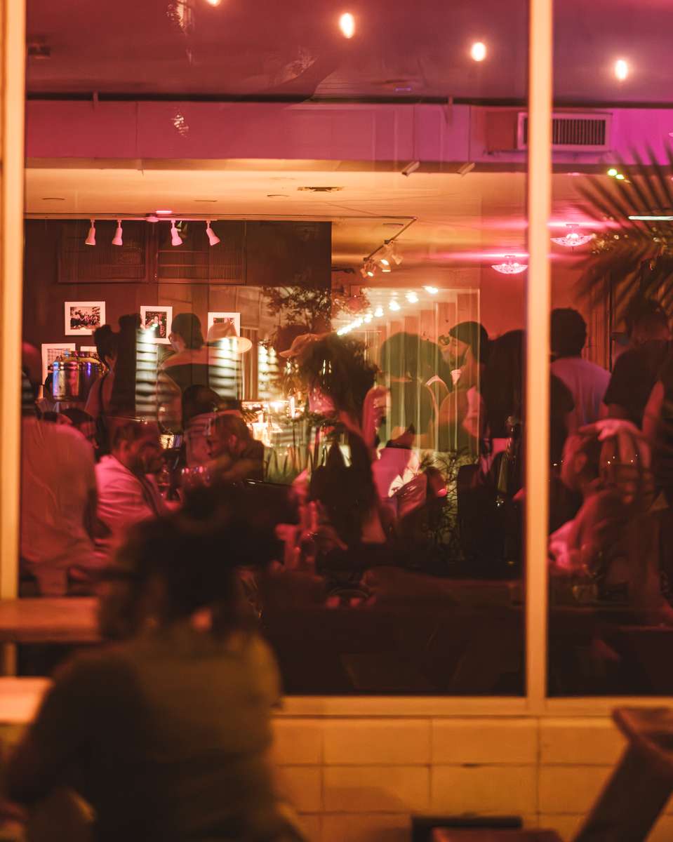 A bustling restaurant scene is viewed through a window, reflecting the warm interior. Patrons are seated at tables, creating a lively, intimate atmosphere.