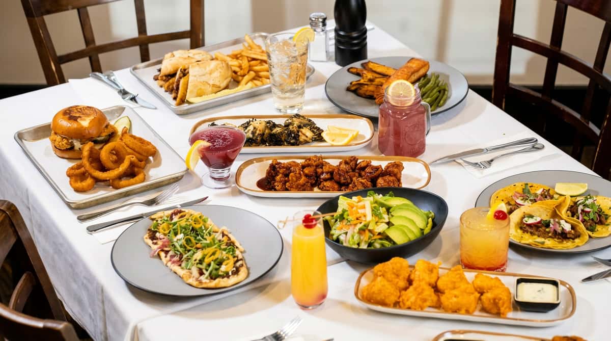 A table laden with various dishes: a burger, fries, onion rings, tacos, flatbread, salads, chicken, and cocktails.