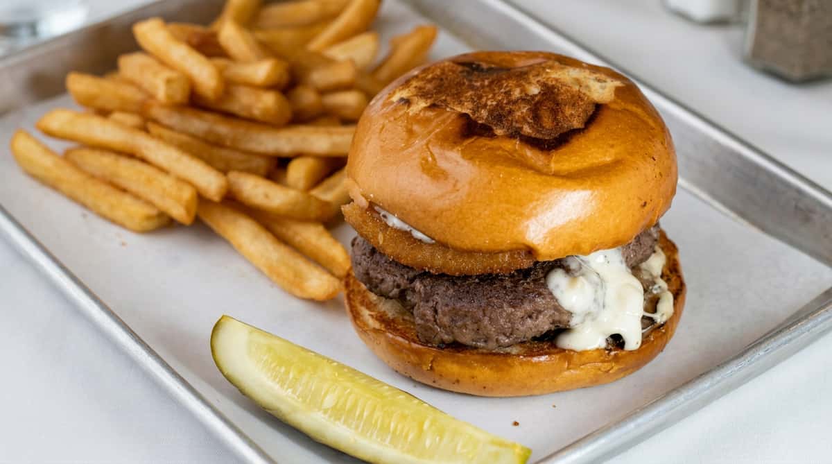 Hamburger with cheese sauce and an onion ring, served with fries and a pickle spear on a metal tray.