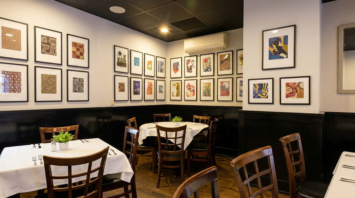 Restaurant dining room with white walls, dark wainscoting, framed artwork, and tables with white tablecloths.