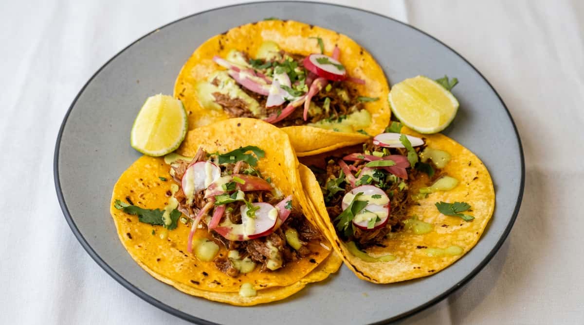 Three street tacos with shredded meat, pickled onions, radishes, cilantro, and green sauce, served with lime wedges.