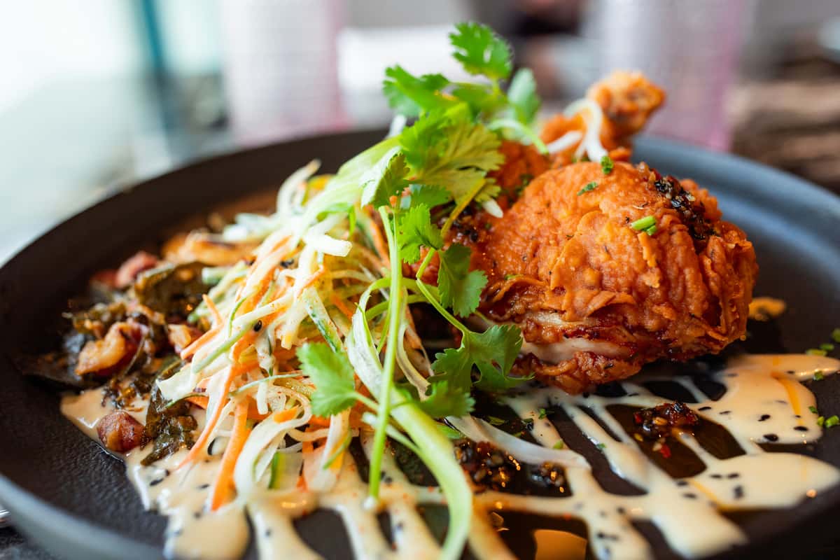 Crispy fried chicken served with slaw, fresh cilantro, and creamy sauce drizzled on a black plate.