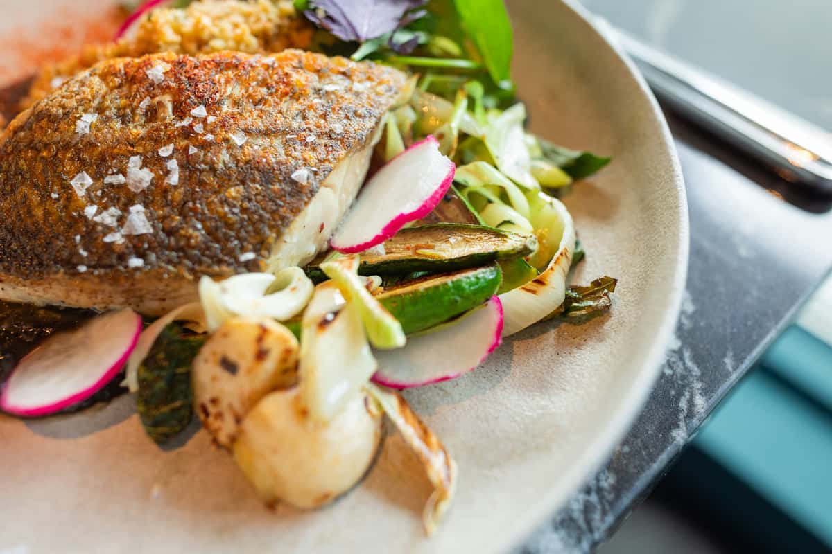 Seared fish fillet with crispy skin served over grilled vegetables, radish slices, and greens on a beige plate.