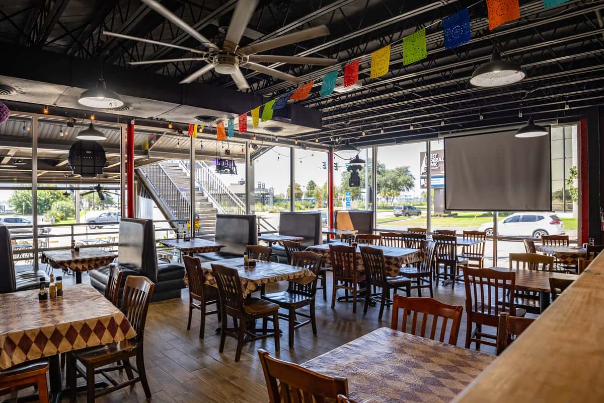 Spacious Mexican restaurant dining area with large windows, papel picado banners, and wooden tables — casual atmosphere with natural light and street-facing views.