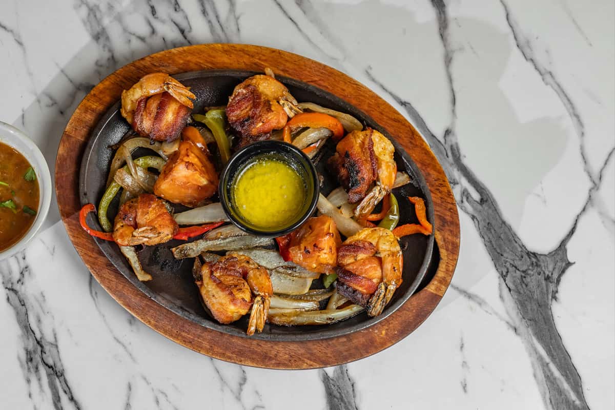 Hot skillet of bacon-wrapped shrimp and grilled vegetables, served with a side of golden garlic butter — sizzling Tex-Mex appetizer known for its smoky flavor and savory presentation.
