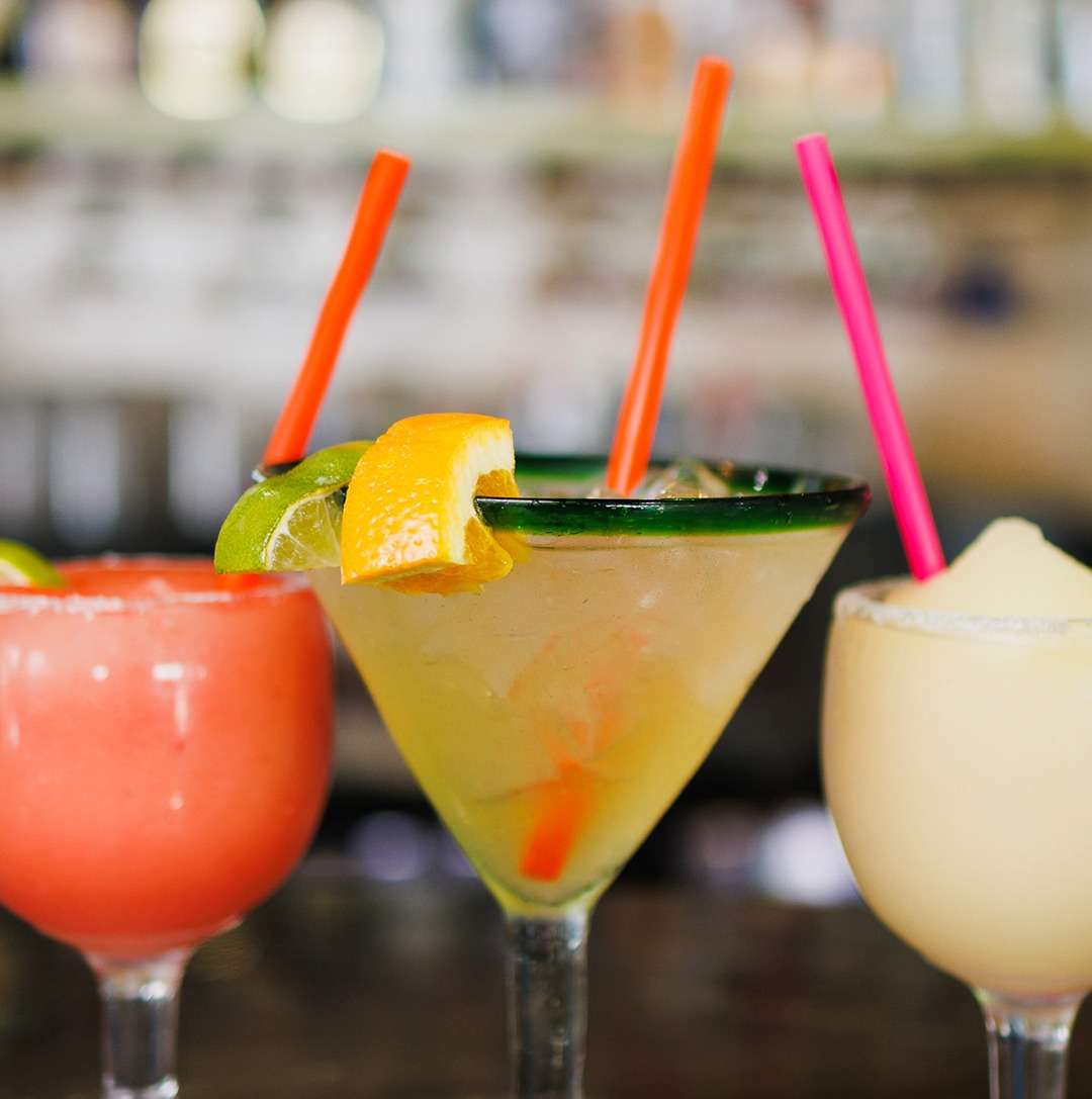 Trio of vibrant margaritas—strawberry, classic lime with orange garnish, and frozen—served in salt-rimmed glasses with colorful straws.