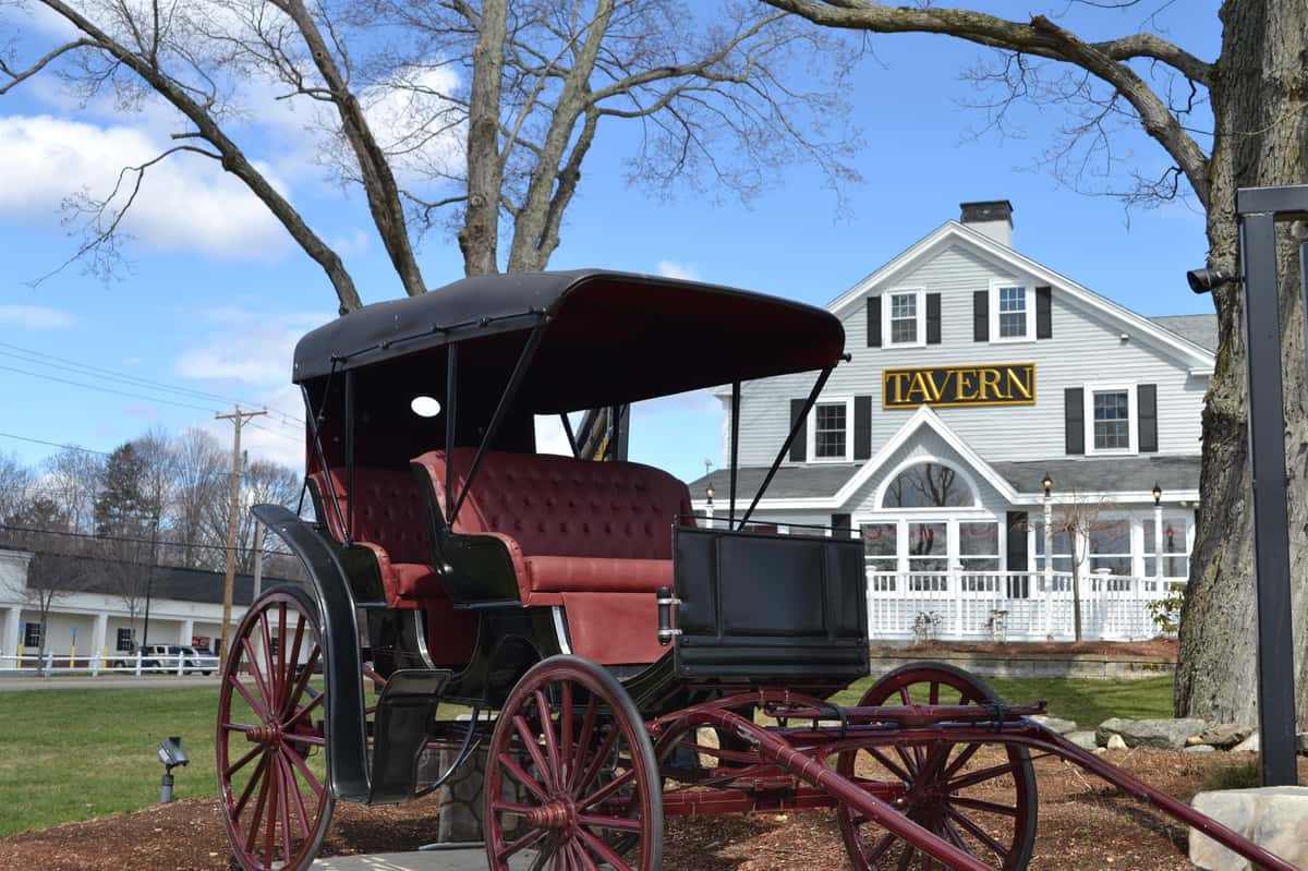 Exterior of White paneled building with "Tavern" sign in middle next to old wooden carriage