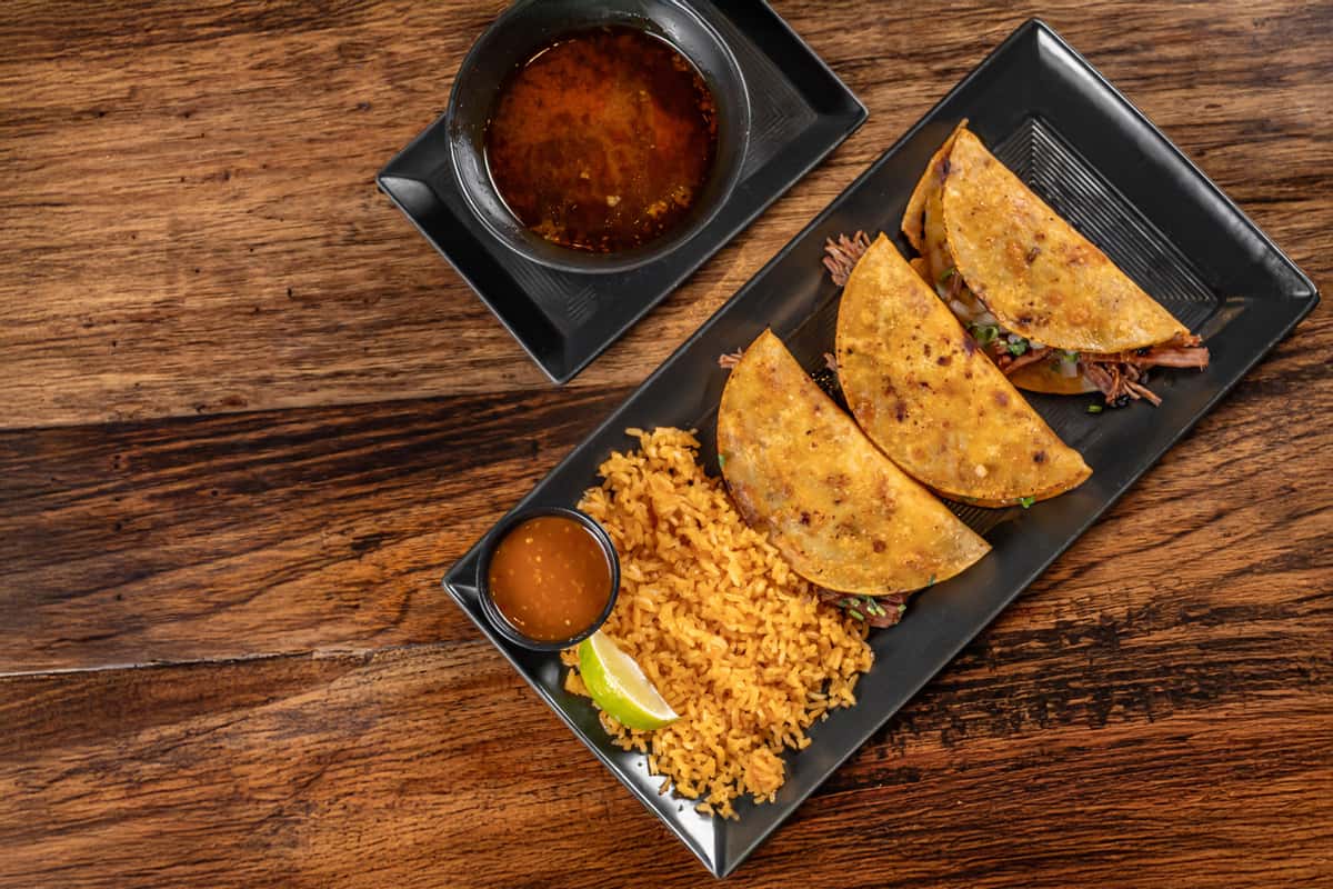 Three golden birria tacos filled with slow-cooked shredded beef, served with Mexican rice, lime wedge, salsa, and a rich consommé dipping broth on a rectangular black plate over rustic wood.