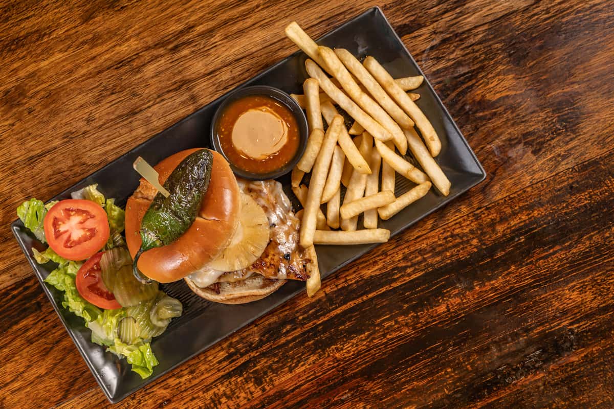 Grilled chicken sandwich topped with melted cheese, grilled pineapple, and a roasted jalapeño on a toasted bun, served with golden French fries, dipping sauce, and a fresh side salad with tomato and pickles.