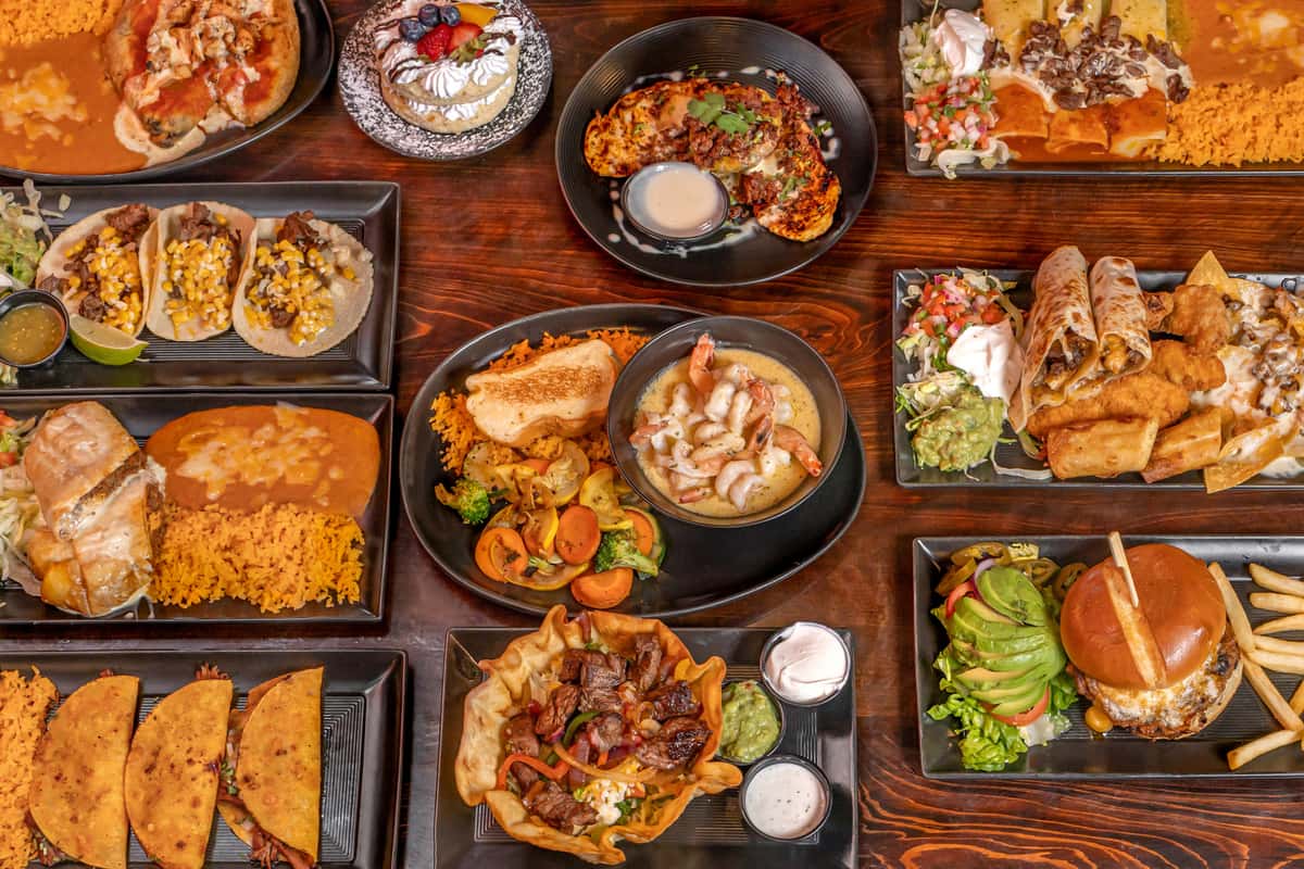 Colorful spread of authentic Mexican cuisine on a rustic wooden table, featuring carne asada tacos topped with corn, shrimp in creamy sauce with grilled vegetables and rice, enchiladas smothered in red sauce, crispy taquitos, quesadillas, a burrito with Spanish rice, taco salad in a fried tortilla shell, and a cheeseburger with avocado and fries. A dessert with whipped cream and fresh fruit is also included.