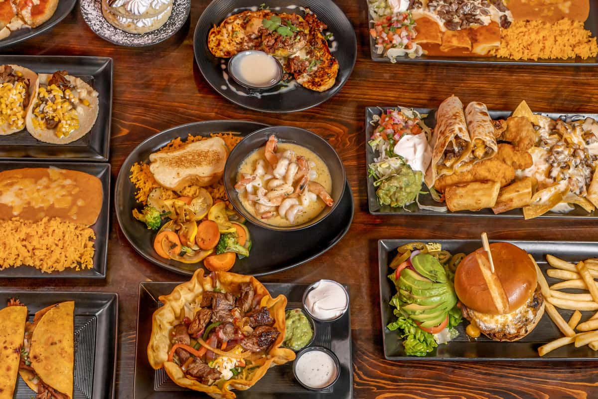 Vibrant assortment of Tex-Mex and Mexican-American dishes including birria tacos, shrimp in creamy garlic sauce, carne asada taco salad, enchiladas with rice and beans, a grilled chicken burger with avocado and fries, crispy taquitos, nachos, and a fresh fruit-topped dessert—served on dark plates over a rustic wood table.