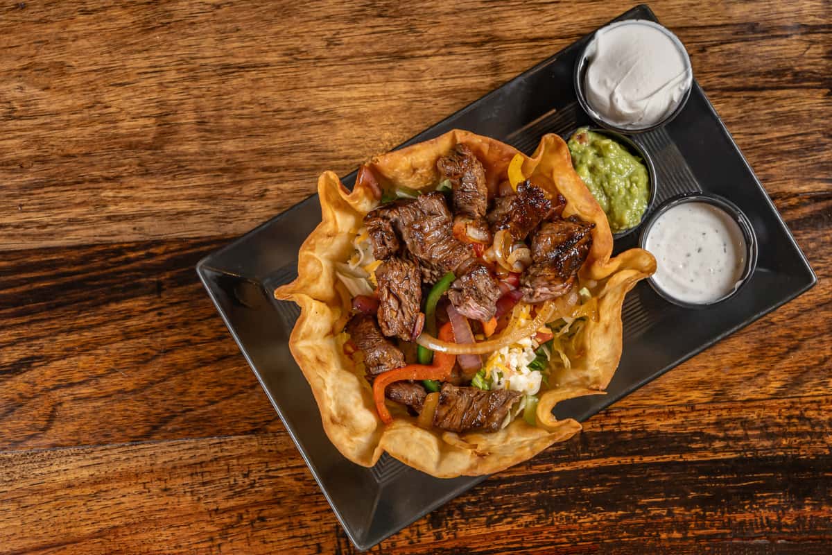 Crispy tortilla taco salad bowl filled with grilled steak, sautéed bell peppers and onions, shredded cheese, and fresh lettuce, served with sides of sour cream, guacamole, and ranch dressing on a sleek black plate over rustic wood.