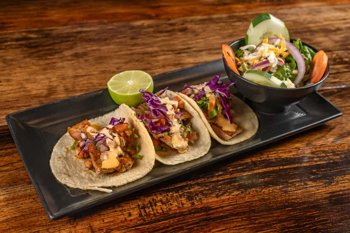 Three grilled fish tacos topped with shredded red cabbage, pico de gallo, and creamy chipotle sauce, served with a lime wedge and a side salad of lettuce, tomato, cucumber, and shredded cheese on a black rectangular plate.