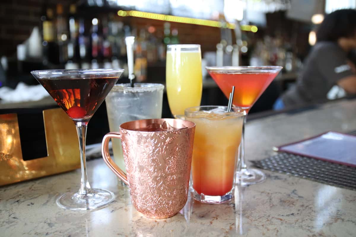 The image shows a neatly arranged selection of colorful cocktails on a bar or tabletop, each in a different style of glass that highlights its unique presentation. There's a copper mug—likely for a mule-style drink—alongside elegant martini and wine glasses, and a tall glass with a layered orange-to-yellow gradient suggesting a tropical or citrus-based cocktail. The drinks range in color from deep red to bright orange and pale yellow, creating a visually appealing spread that feels both refreshing and upscale. The overall presentation suggests a curated cocktail menu, ideal for a stylish dining or social setting.
