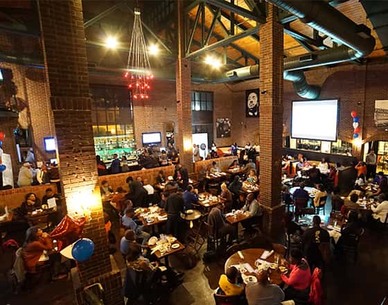 The image shows a vibrant, upscale restaurant interior with a lively, social atmosphere, likely taken during a busy evening service. The space features high ceilings, warm ambient lighting, and a modern-industrial design with large windows reflecting city lights. Tables are closely arranged and filled with guests, creating an energetic, bustling feel, while soft candlelight adds a touch of intimacy. In the background, there appears to be a screen or stage area, suggesting the venue may host events or live entertainment. Overall, the setting feels stylish, crowded, and ideal for a dynamic dining or social experience rather than a quiet meal.