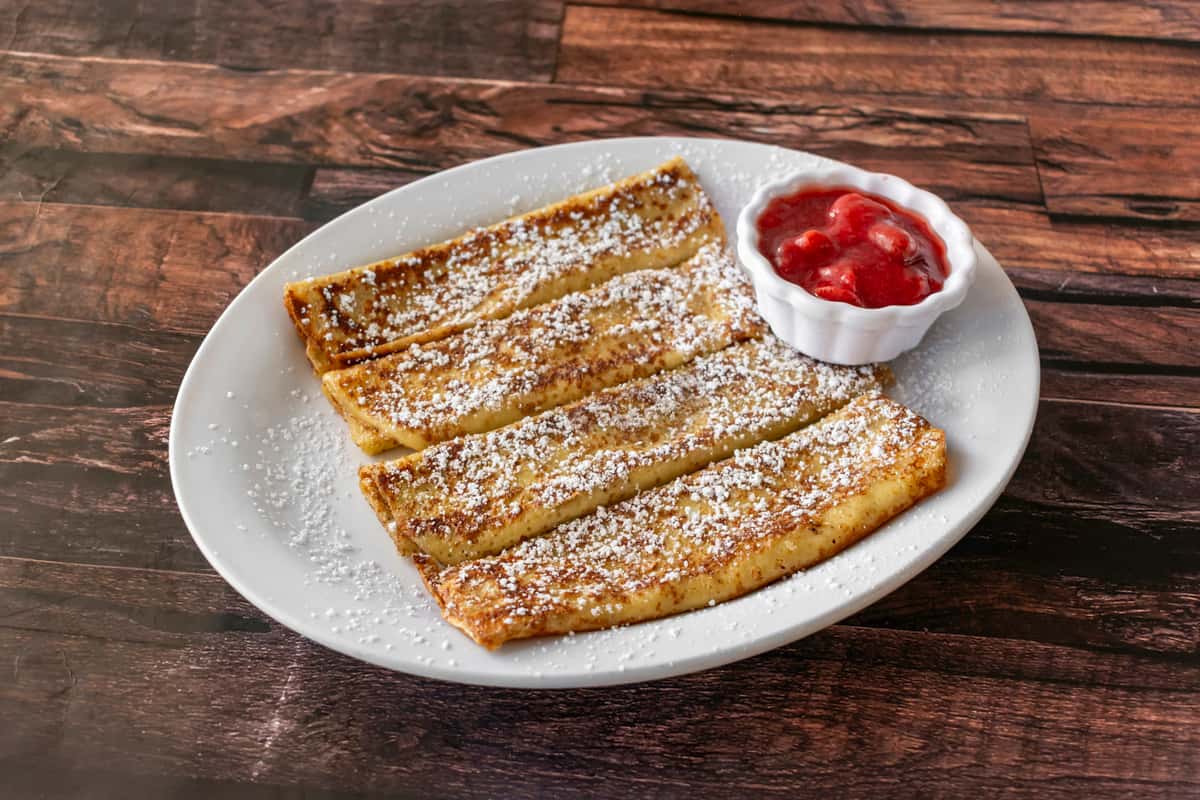 Sliced French toast sticks dusted with powdered sugar, served with a side of strawberry compote on a white plate over a rustic wood table — a kid-friendly breakfast option ideal for diners or casual brunch menus.