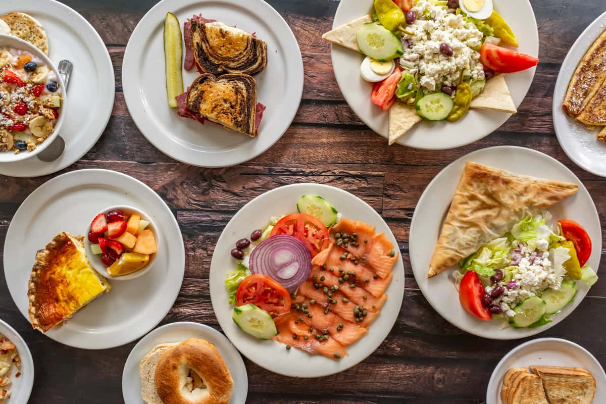 Overhead view of assorted breakfast and lunch dishes including quiche, smoked salmon with veggies, Greek salad with pita, a Reuben sandwich, yogurt parfait, and fresh fruit — a diverse and inviting selection from a daytime diner or brunch café.